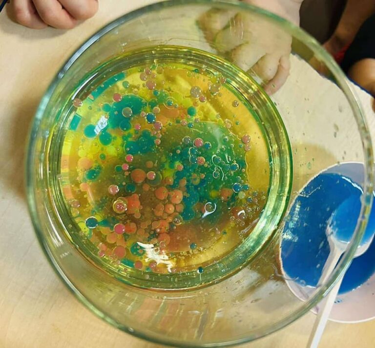 Toddler doing colorful oil and water experiment with food coloring bubbles floating in glass bowl.