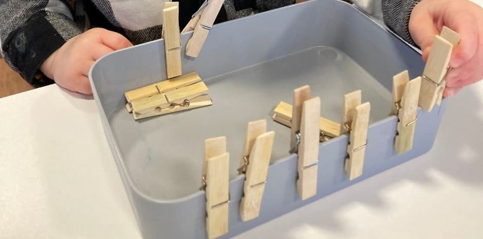 toddler clips clothespins around the edge of a container and experiments with attaching and removing them.